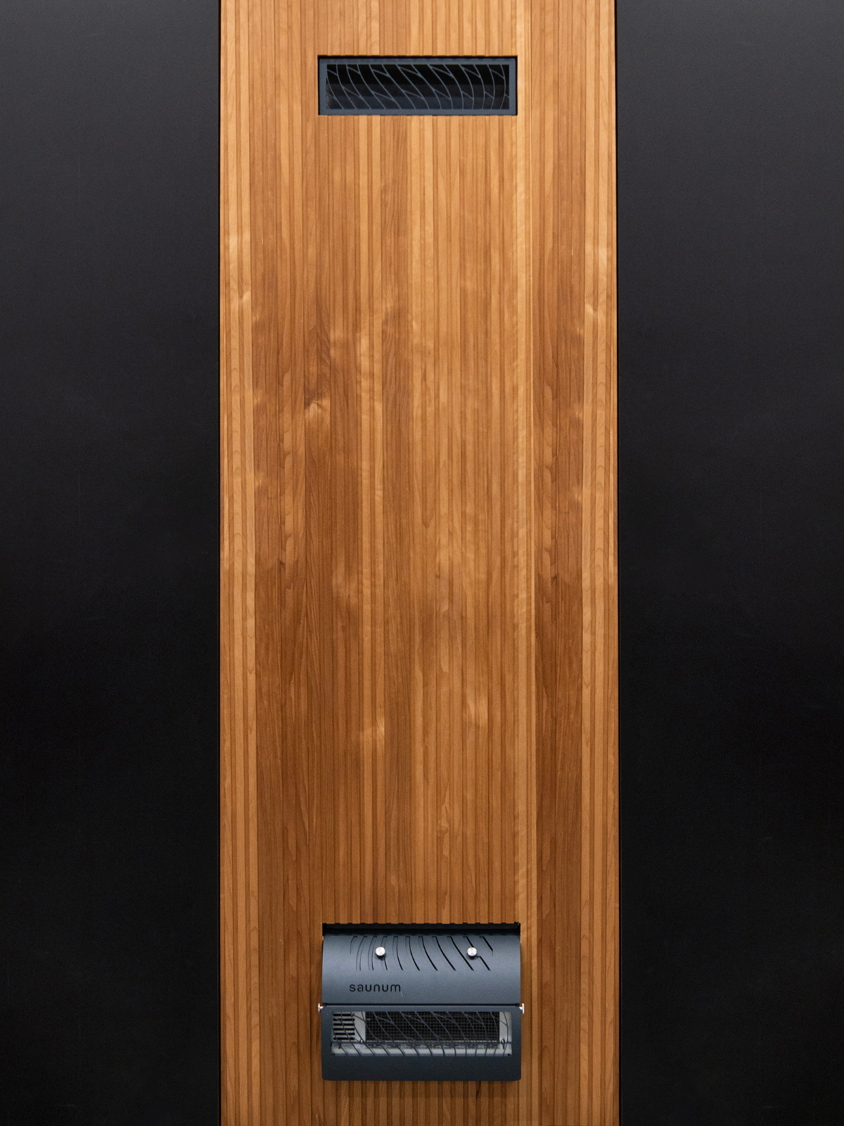 Modern sauna heater with a sleek, dark metal cover displays a grill design, set against light wooden panels and a rack of sauna stones. Cozy and warm ambiance.