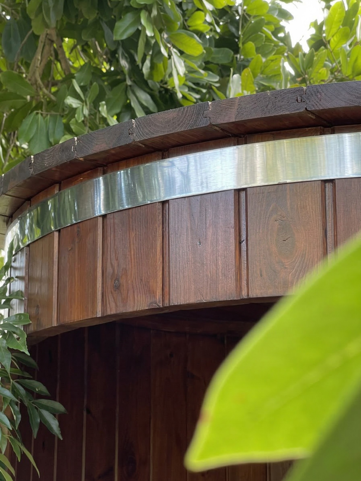 Wooden hot tub with metallic rim surrounded by green foliage
