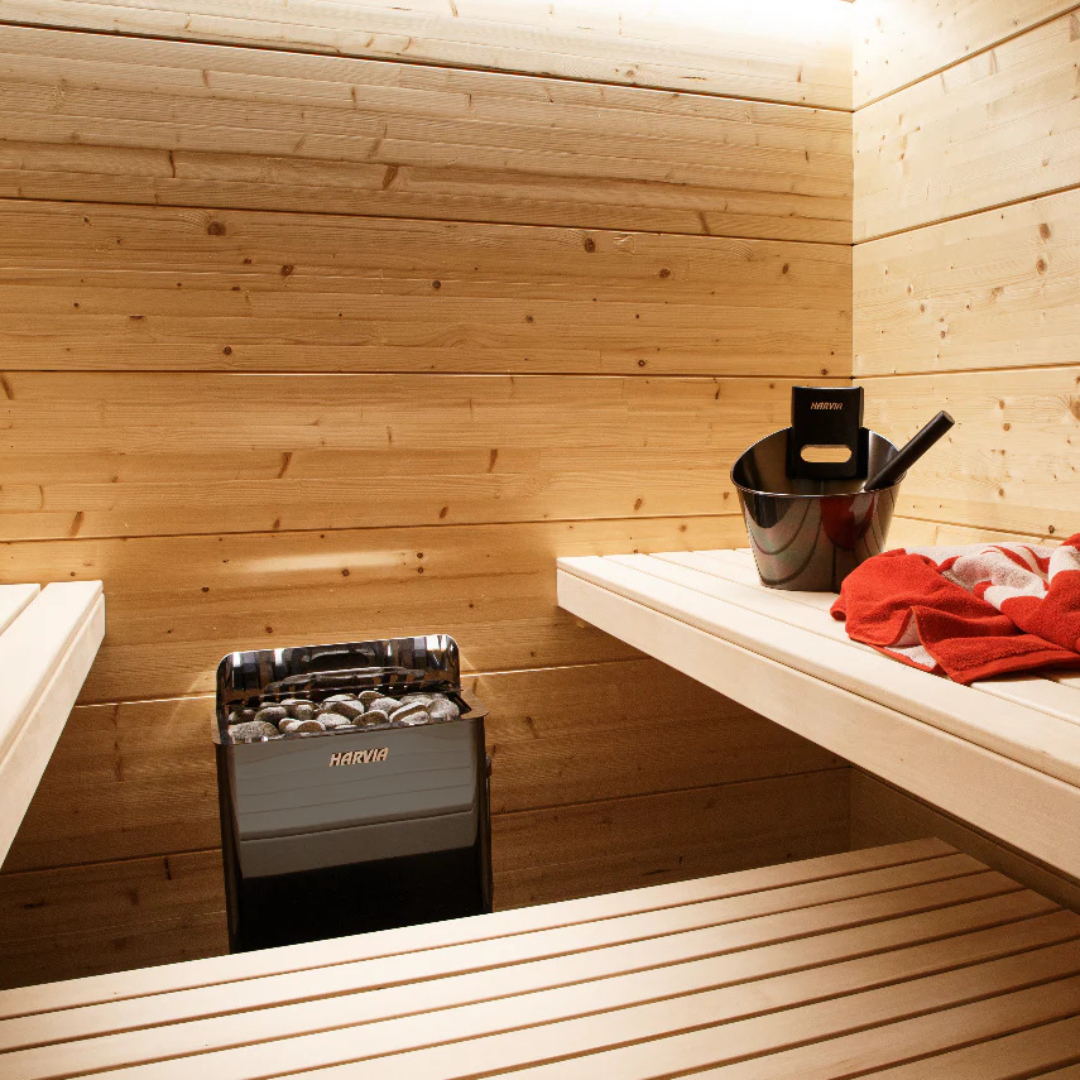 Wooden sauna interior with a central stove filled with stones. Benches line the walls, with a black bucket and ladle, and a red towel on the right. Calm atmosphere.