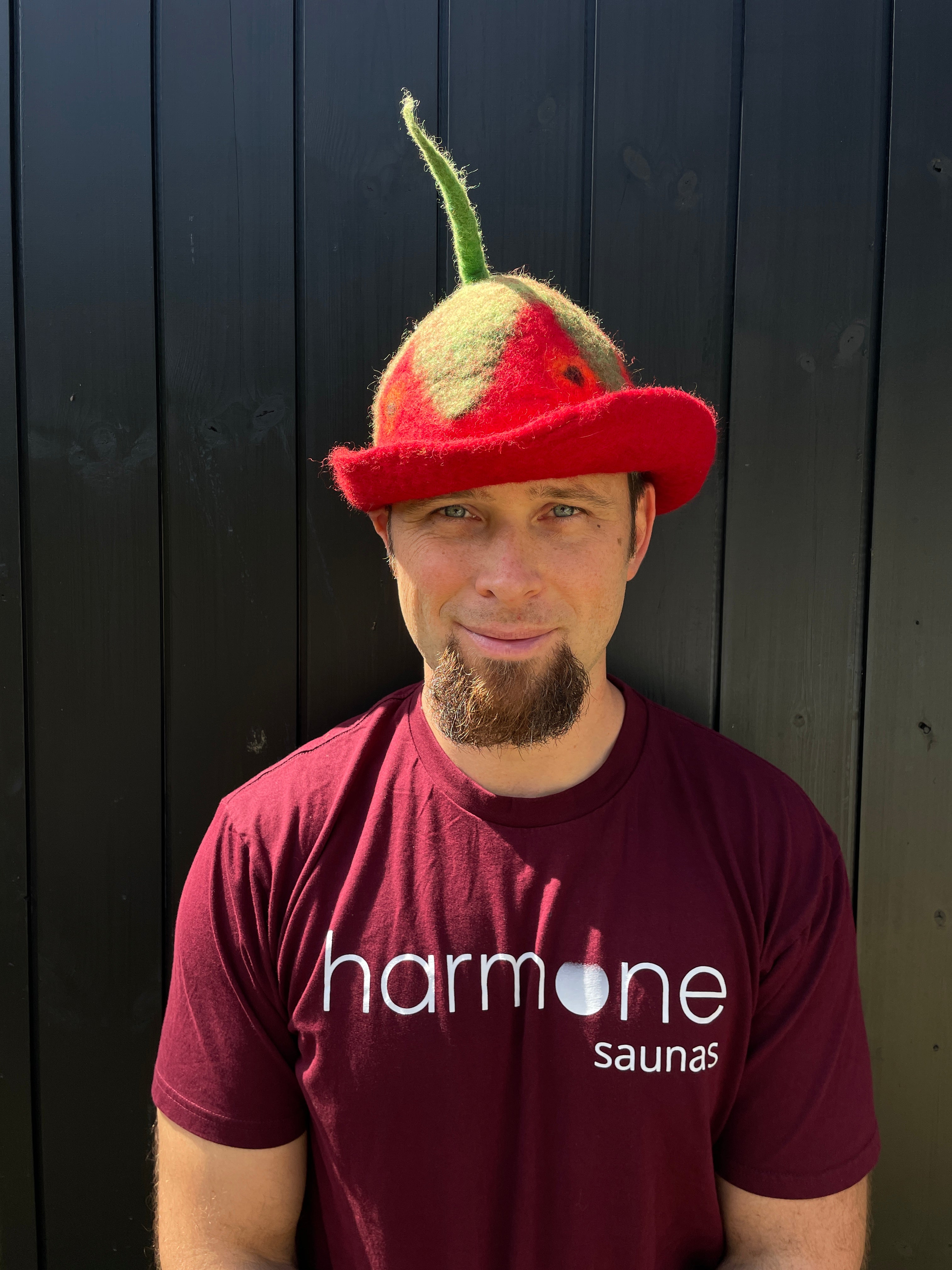 Person wearing a red cap with a green brim and a maroon shirt with 'harmone saunas' logo against a dark wooden background.