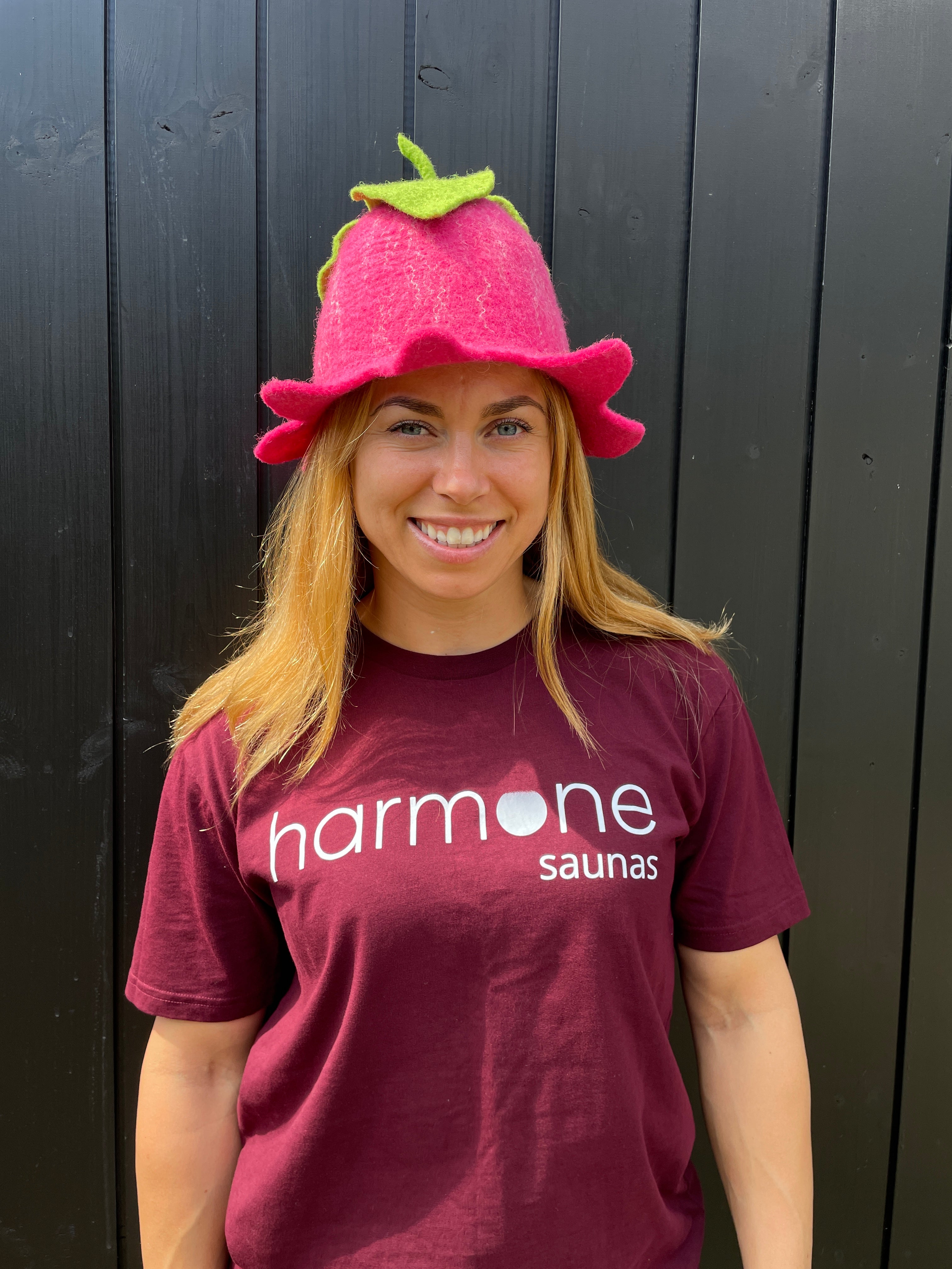 Person wearing a maroon 'harmone saunas' shirt and pink hat with green stem on a dark wooden background