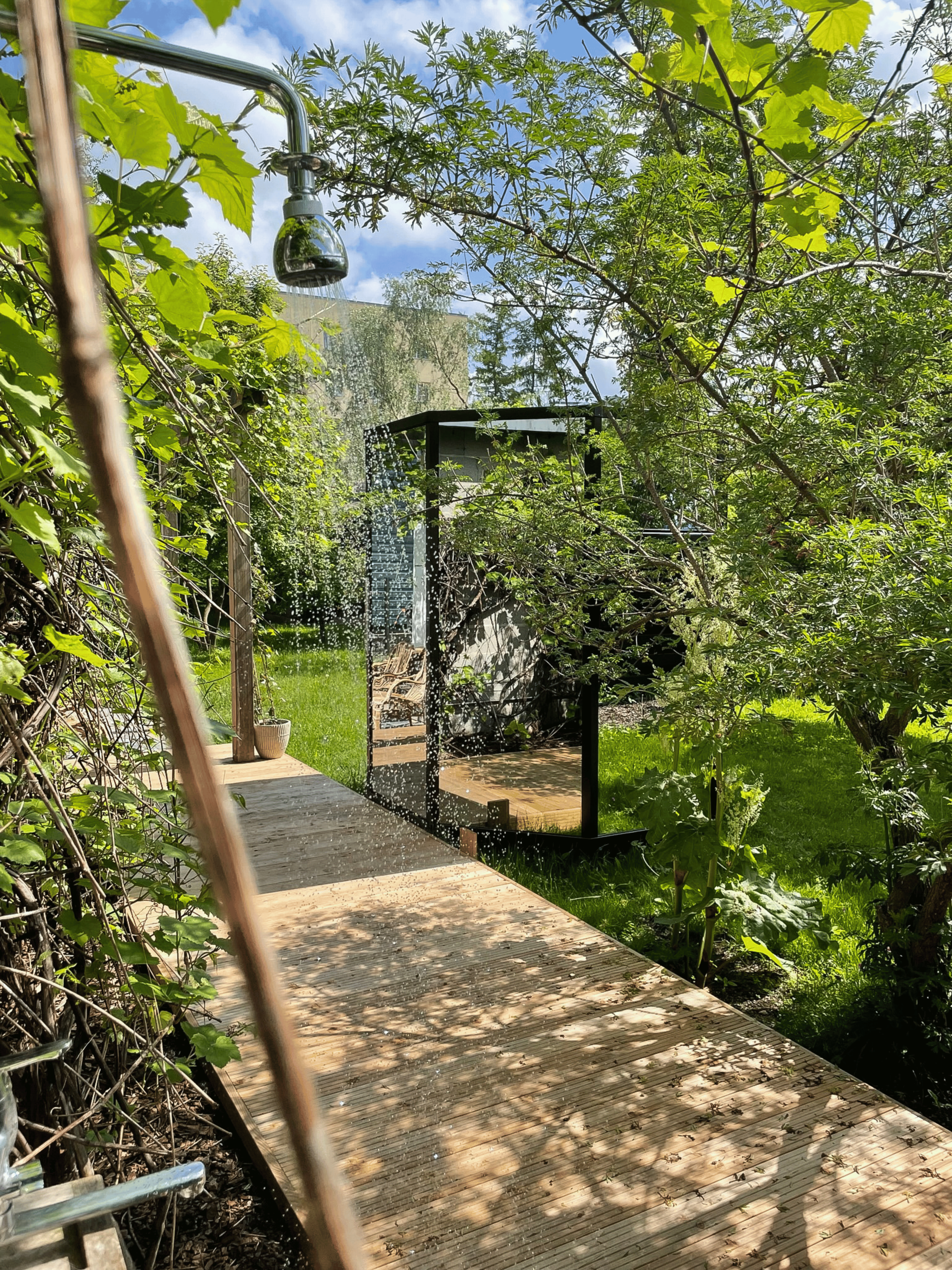 Outdoor shower in a lush green garden with trees and a clear sky.