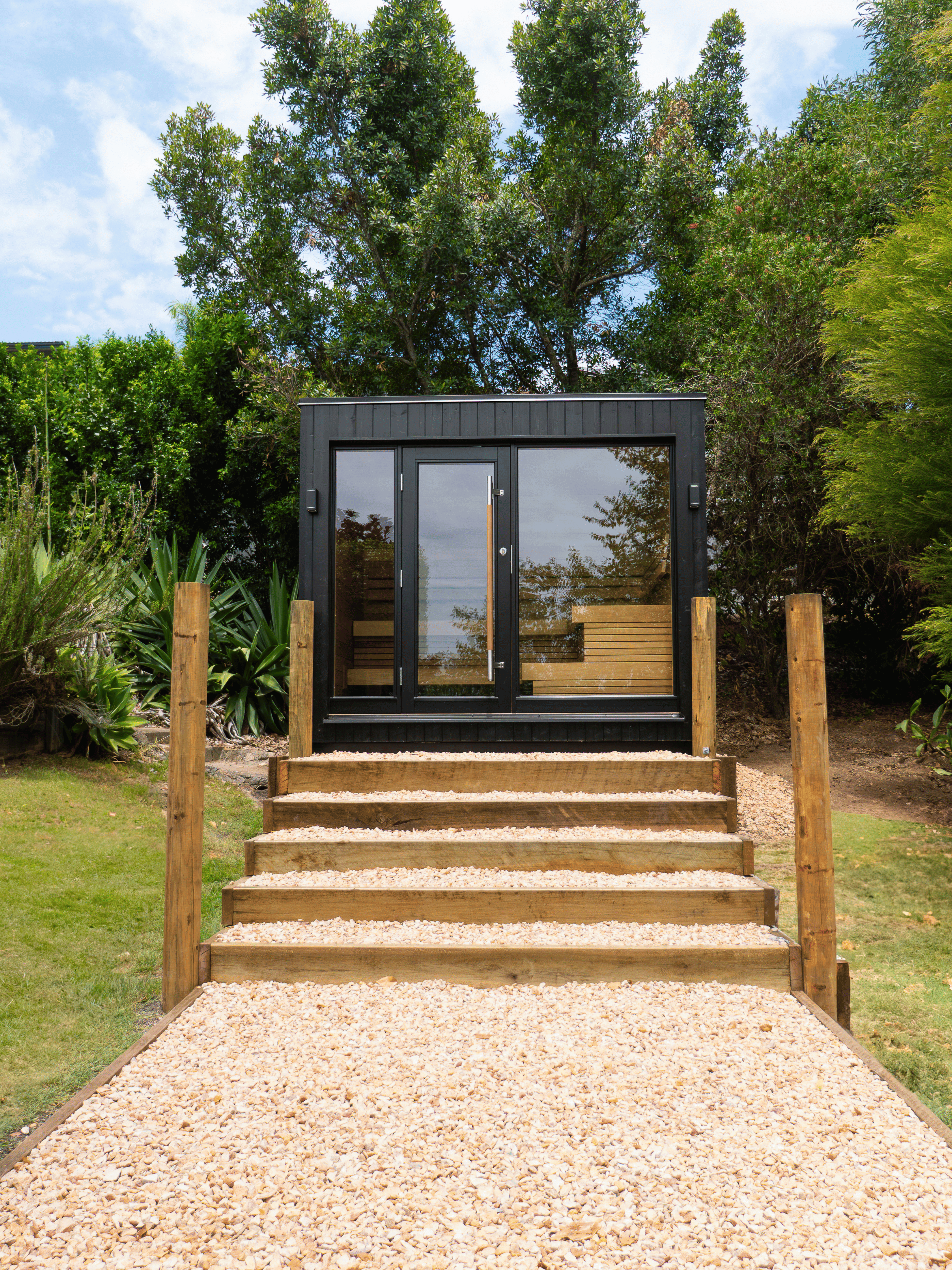 Modern garden office with glass doors and steps leading up to it, surrounded by greenery.