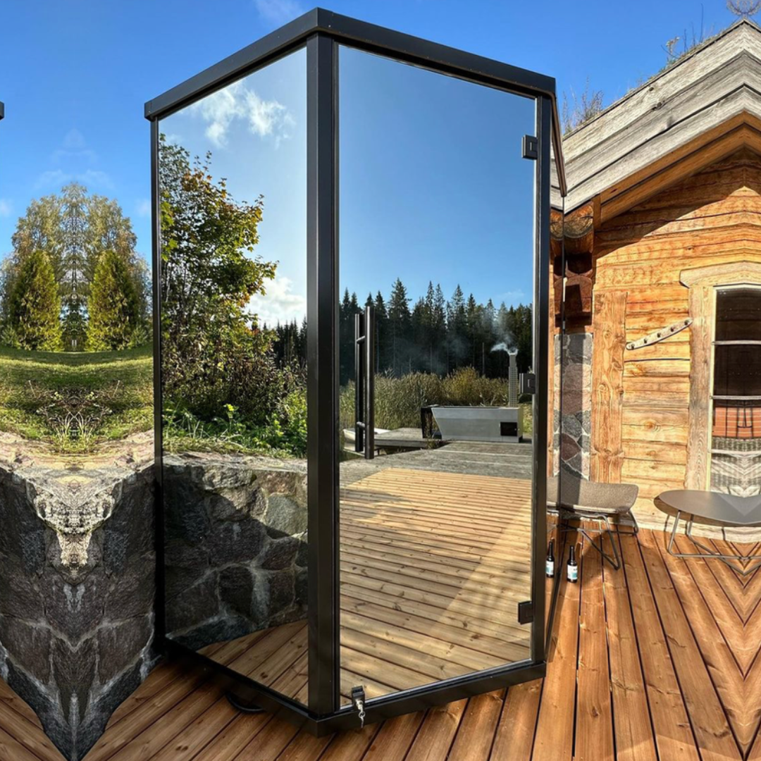 A glass-walled outdoor shower cabin on a wooden deck near a rustic cabin. Reflects trees and blue sky, evoking a tranquil, natural vibe.