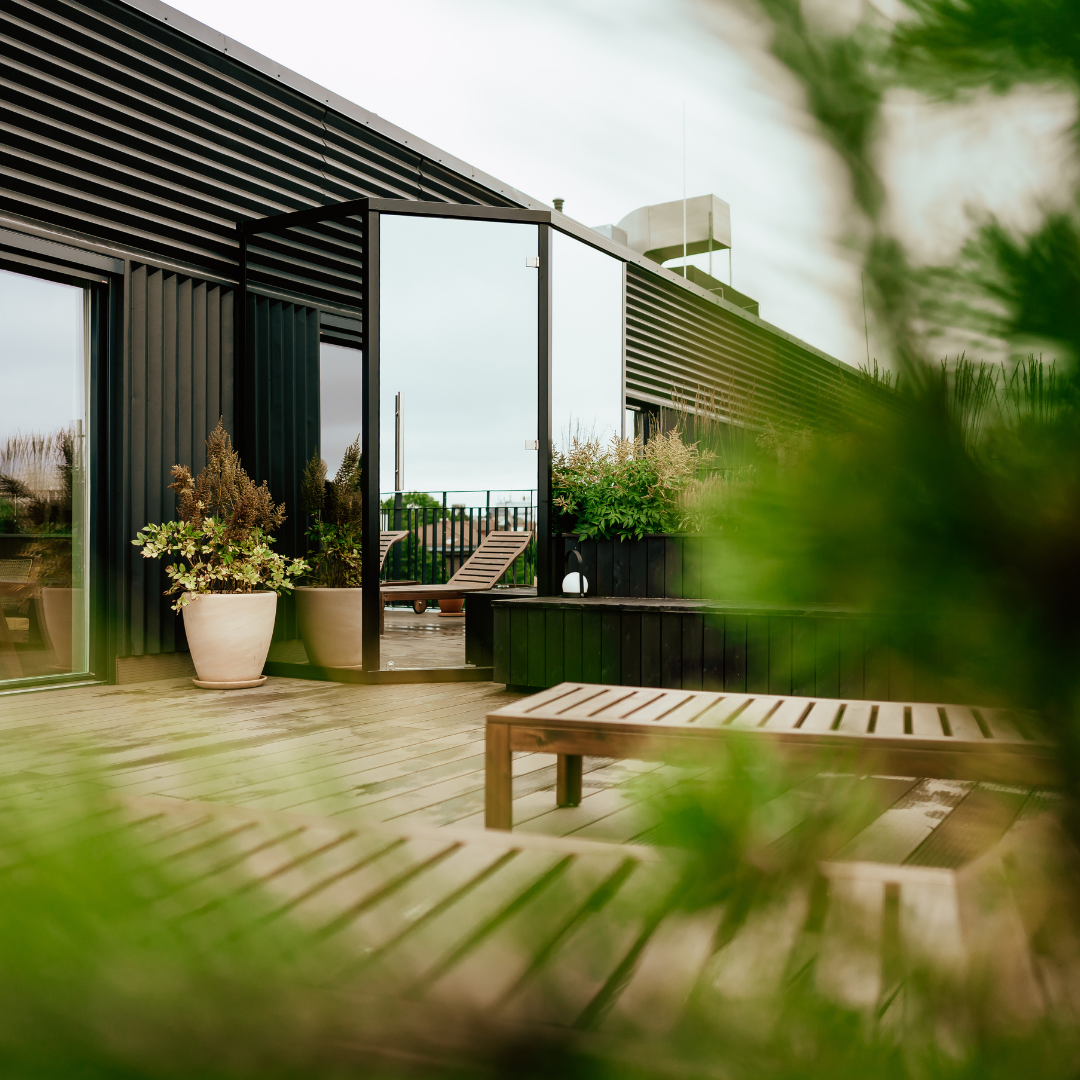 Modern rooftop terrace with wooden benches, planters filled with green foliage, and an open glass door leading indoors. Calm and inviting atmosphere.
