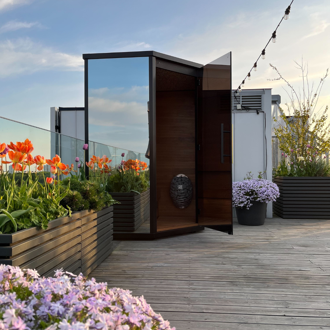 Rooftop garden with vibrant tulips and flowering plants in planters. A modern glass and wood booth is open under string lights against a sunset sky.