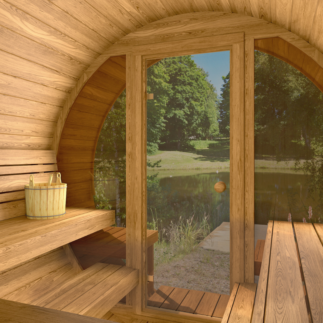 Interior of a wooden sauna with curved walls, featuring a bucket on benches. A glass door offers a view of a serene, tree-lined pond.