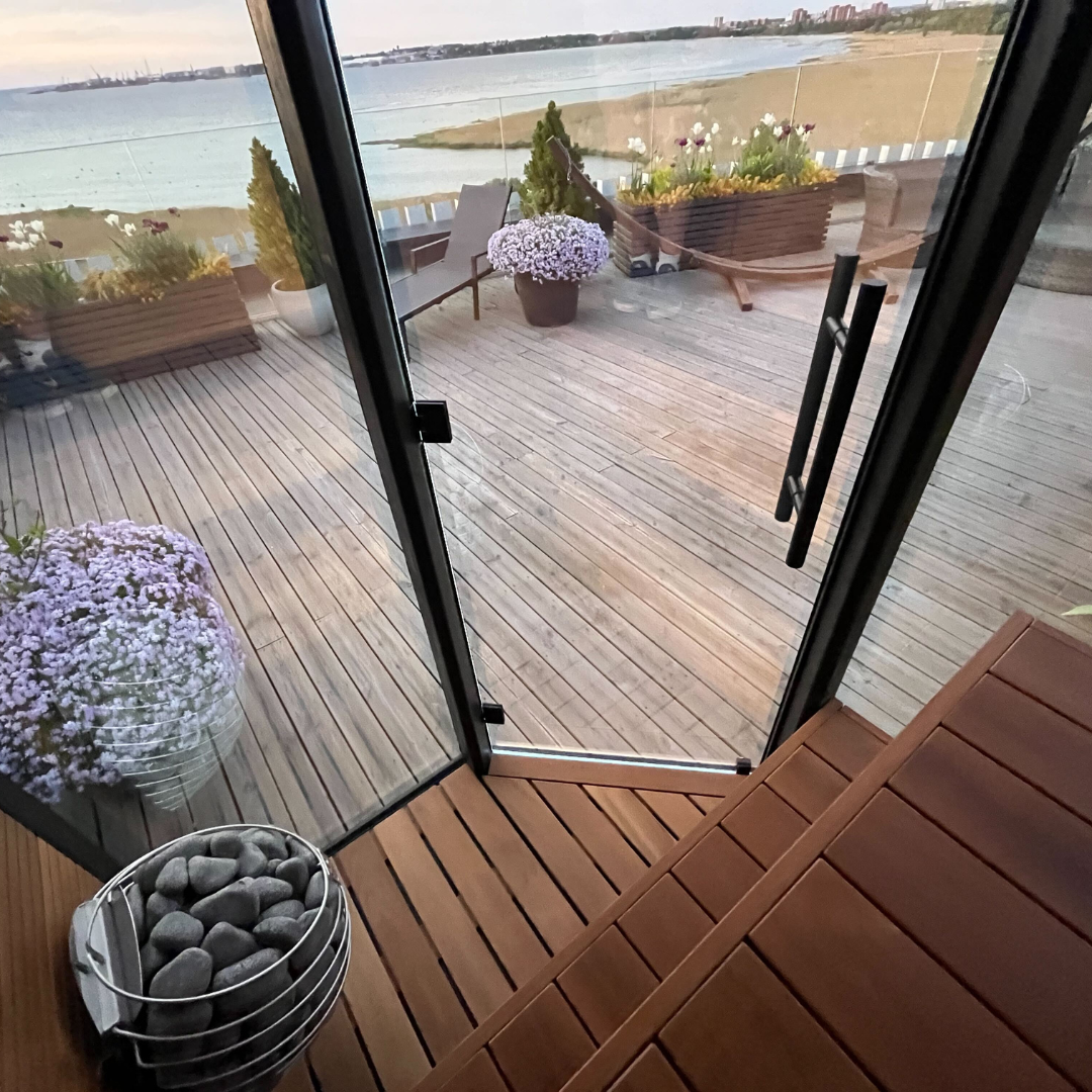 A serene rooftop terrace overlooking a beach, with wooden decking, potted purple flowers, and a glass door, creating a peaceful and inviting atmosphere.