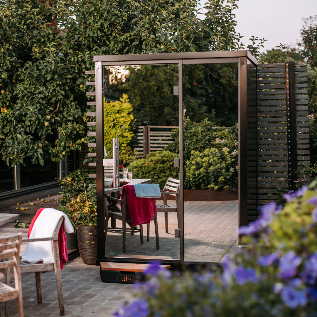 A compact outdoor sauna with a floor-to-ceiling mirror-glass facade, surrounded by greenery. The sauna is made of thermo-alder wood and includes a Harvia 'The Wall' heater.