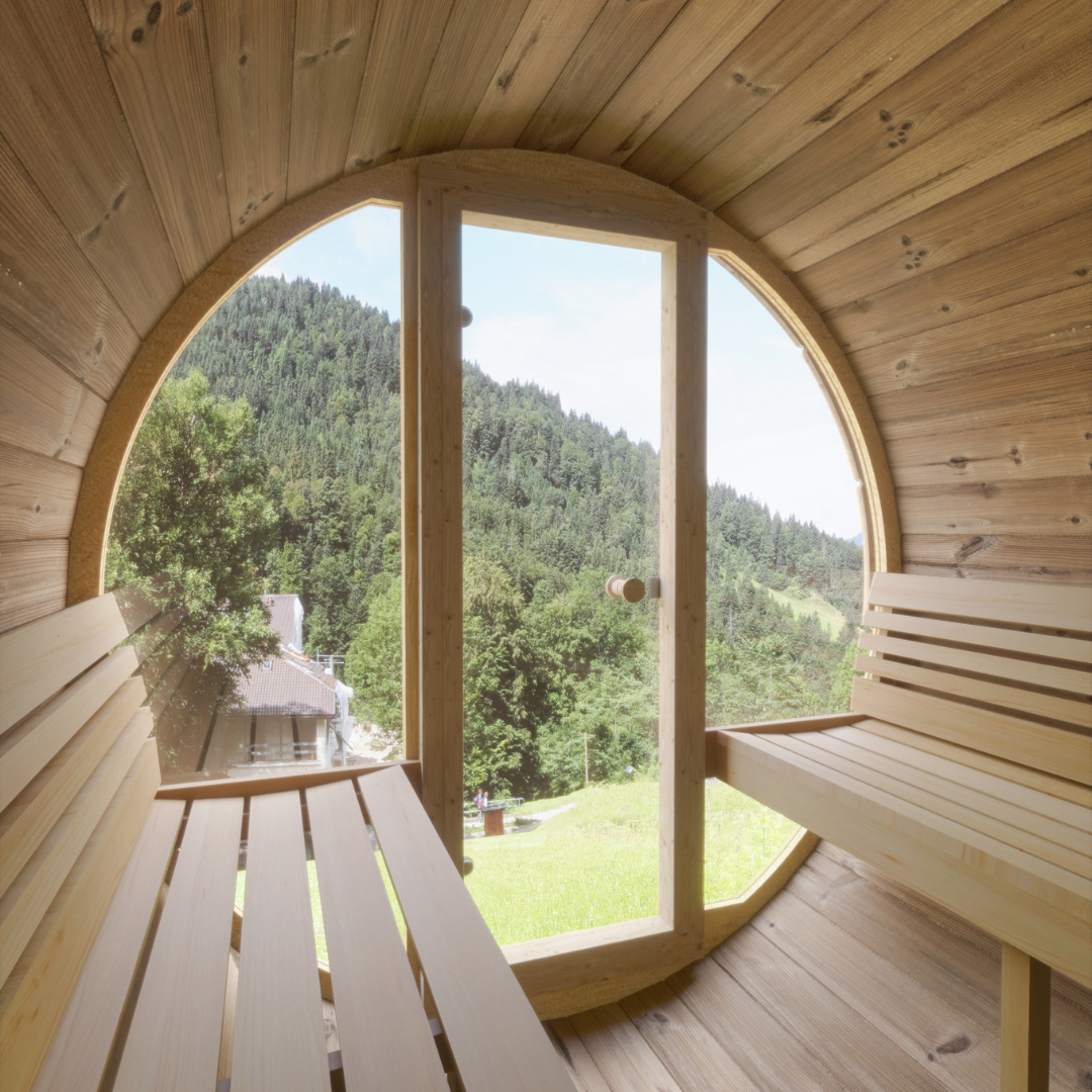 Wooden barrel sauna with benches, featuring a large circular window framing a serene view of lush green mountains and a bright, sunny sky.