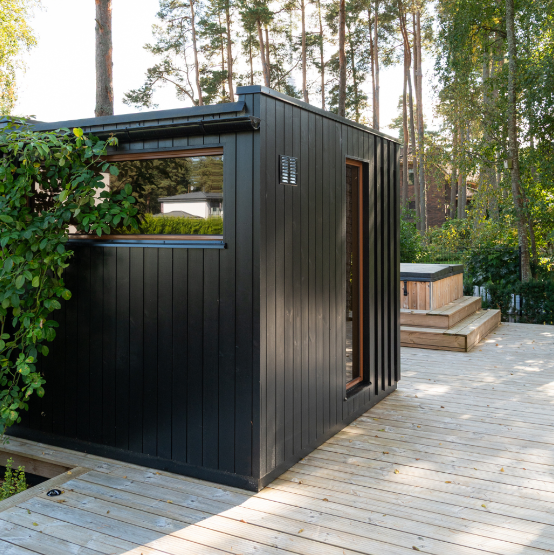 Modern black cabin in a forest setting with wooden deck and greenery.