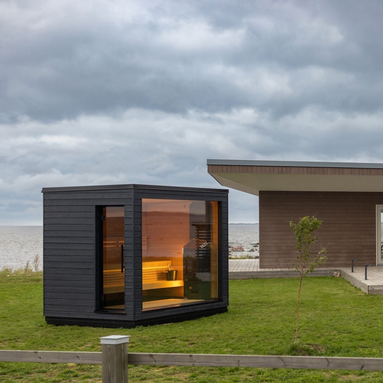 Modern black sauna with large windows sits on lush green grass near a contemporary building. The sky is overcast, creating a serene, calming atmosphere.