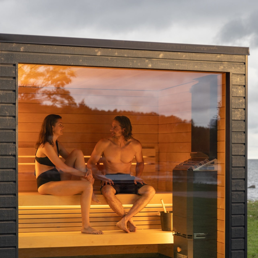 A man and woman relax in a modern, wooden sauna with warm, orange lighting, smiling at each other. It's set against an outdoor backdrop with a cloudy sky.