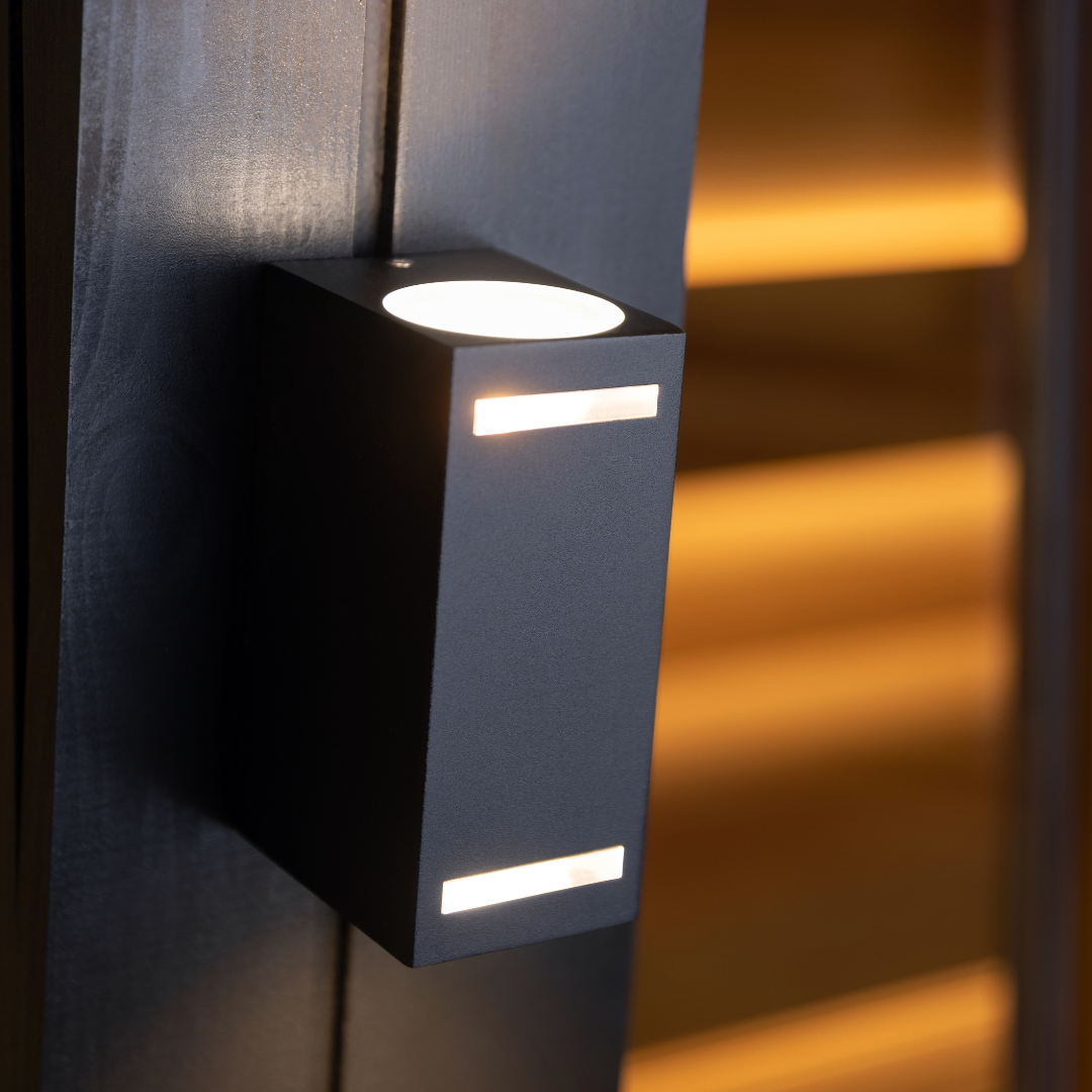 A modern, black outdoor wall light with a minimalist design casts a warm glow. Positioned on a dark surface, it creates a cozy ambiance.