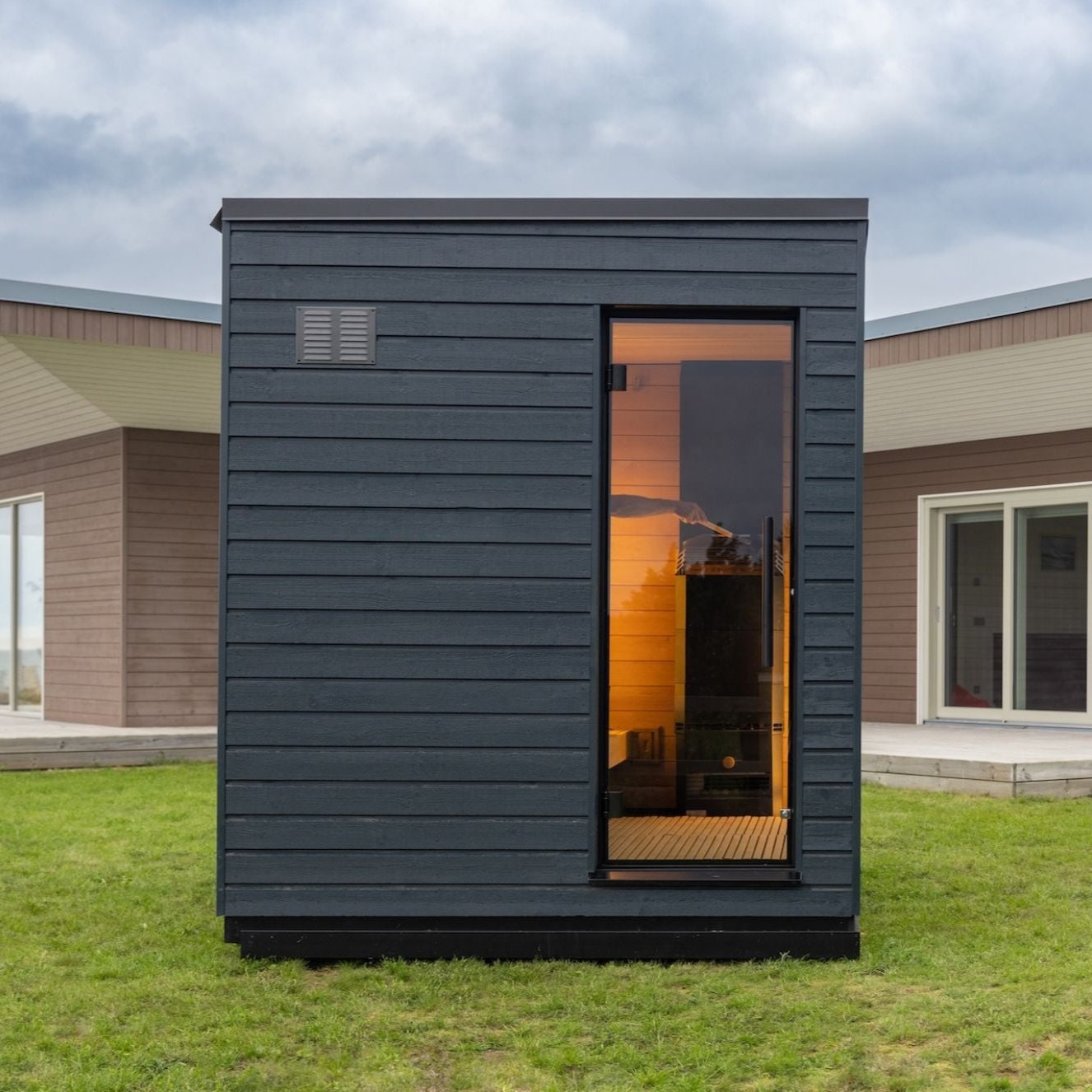 Modern black wooden sauna cabin with a glass door emits a warm glow on a grassy lawn. A larger building is visible in the background under cloudy skies.