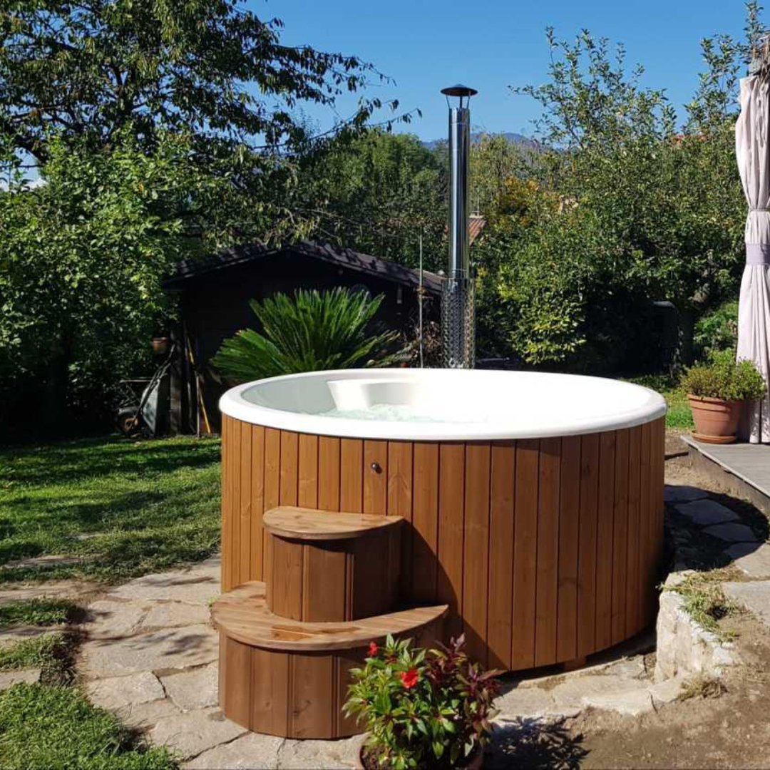 A wooden hot tub with steps sits on a stone patio in a lush garden. Surrounded by trees and plants, it conveys a peaceful, inviting outdoor retreat.