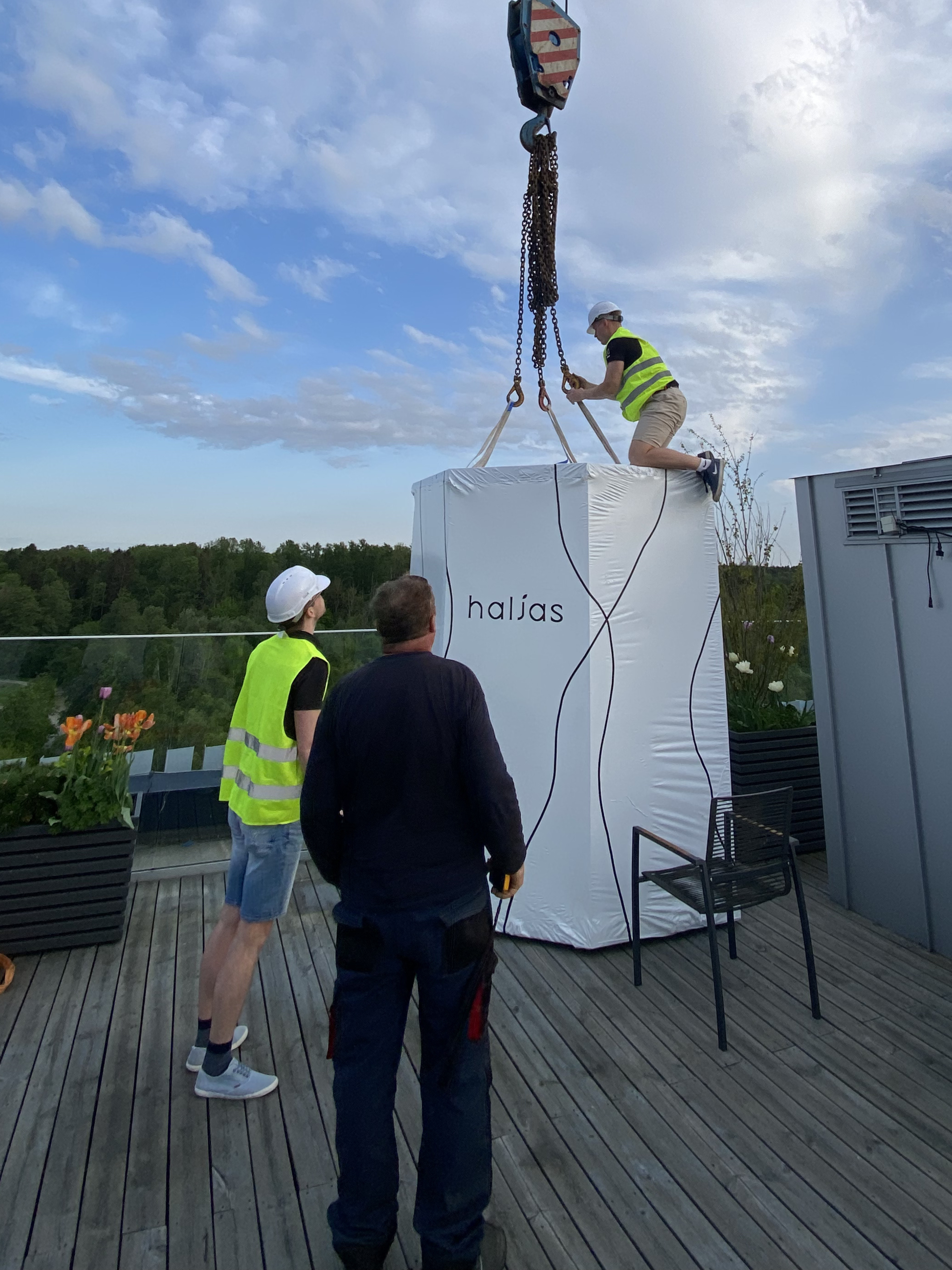 Three people working on a large white container labeled 'halas' with a crane on a rooftop.