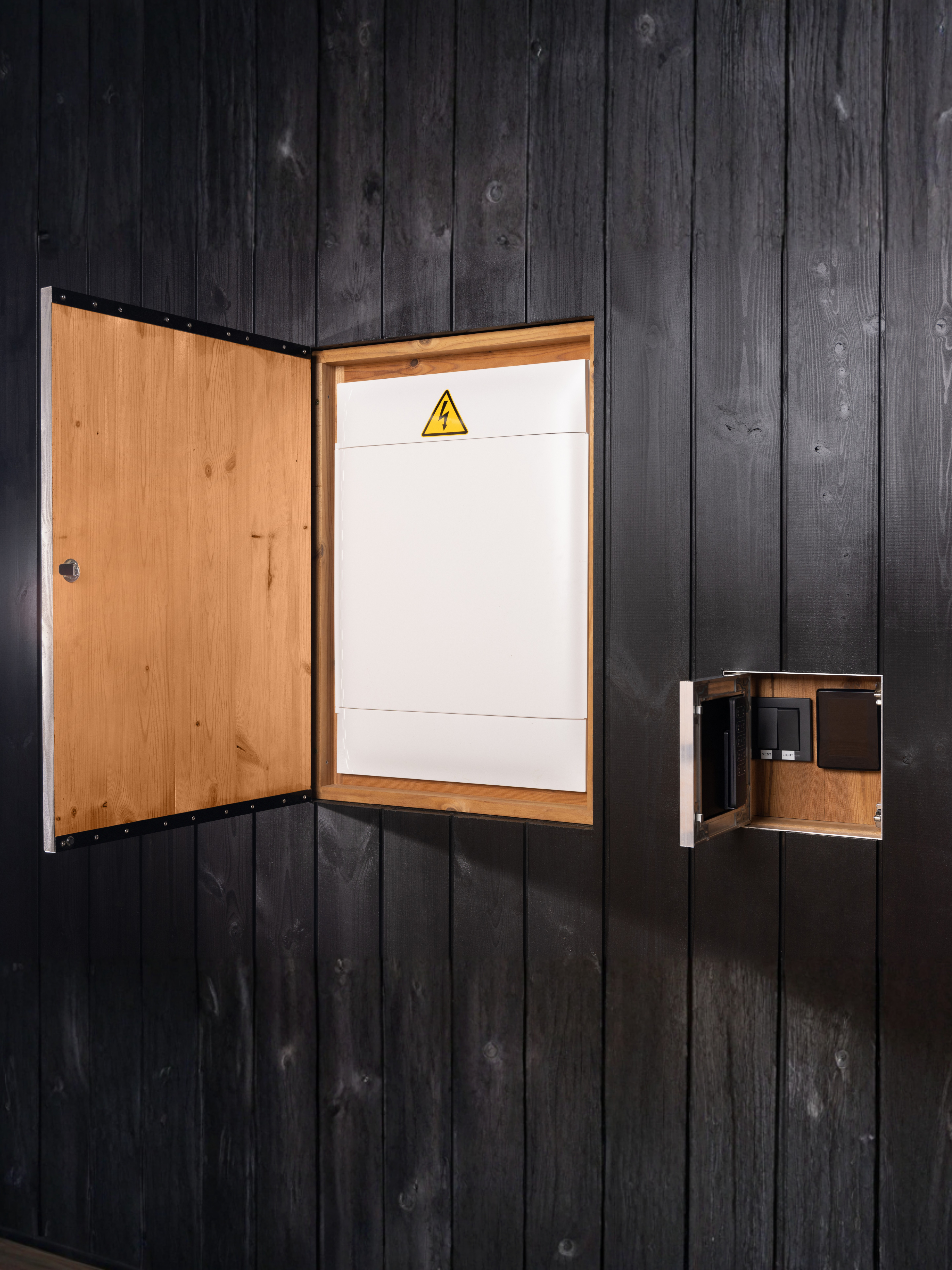 Wooden electrical panel box with a warning label on a black paneled wall.