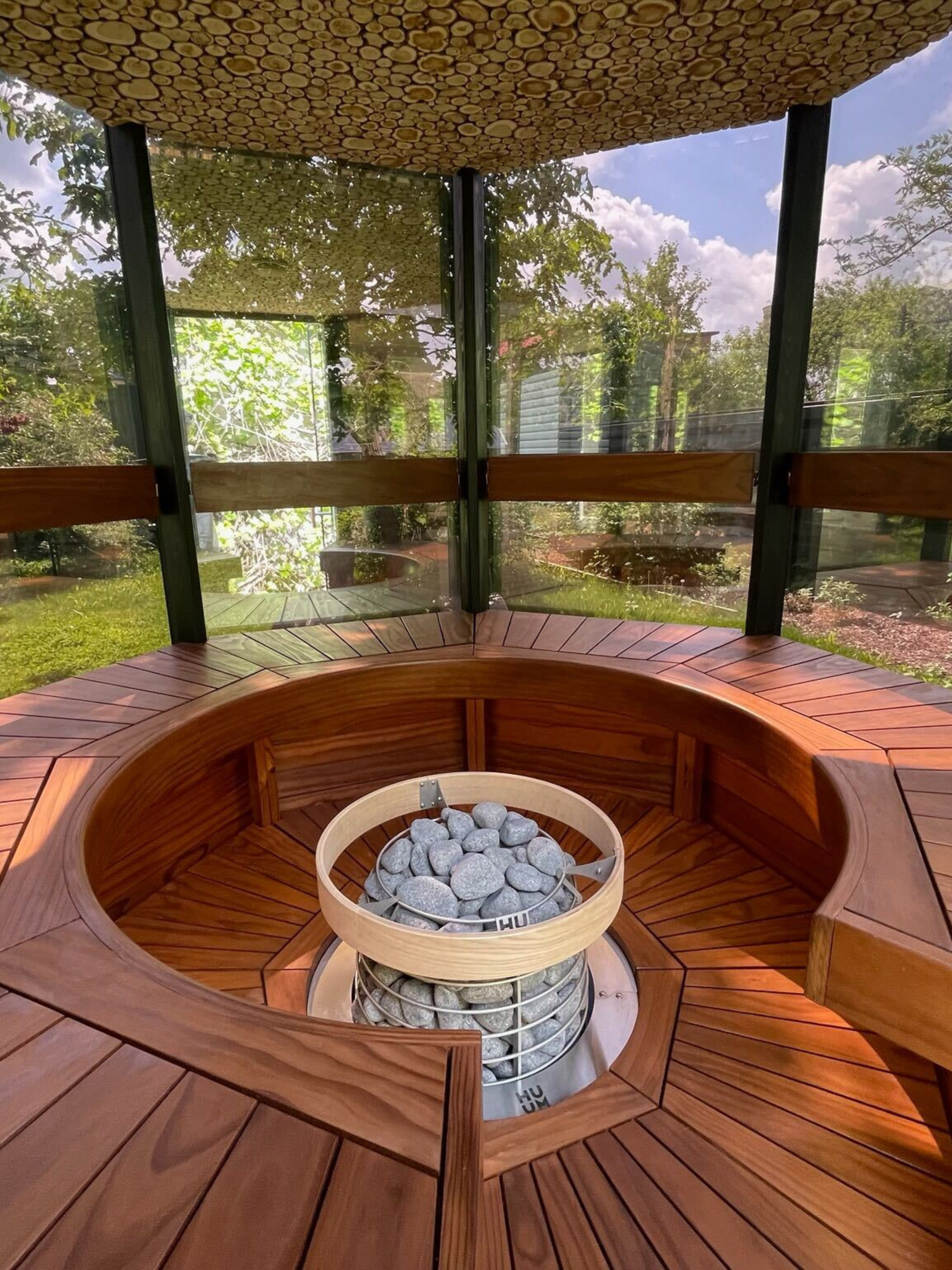 Wooden outdoor structure with a fire pit and surrounding glass walls, set against a natural background.