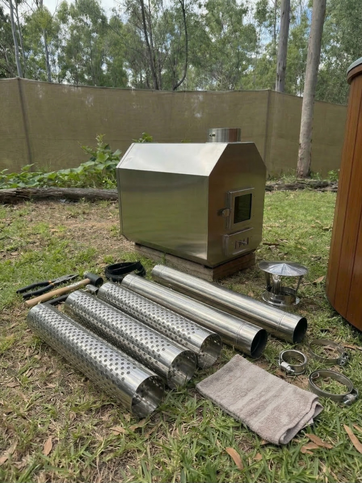 Stainless steel barbecue grill and accessories on grass with a wooden fence and trees in the background