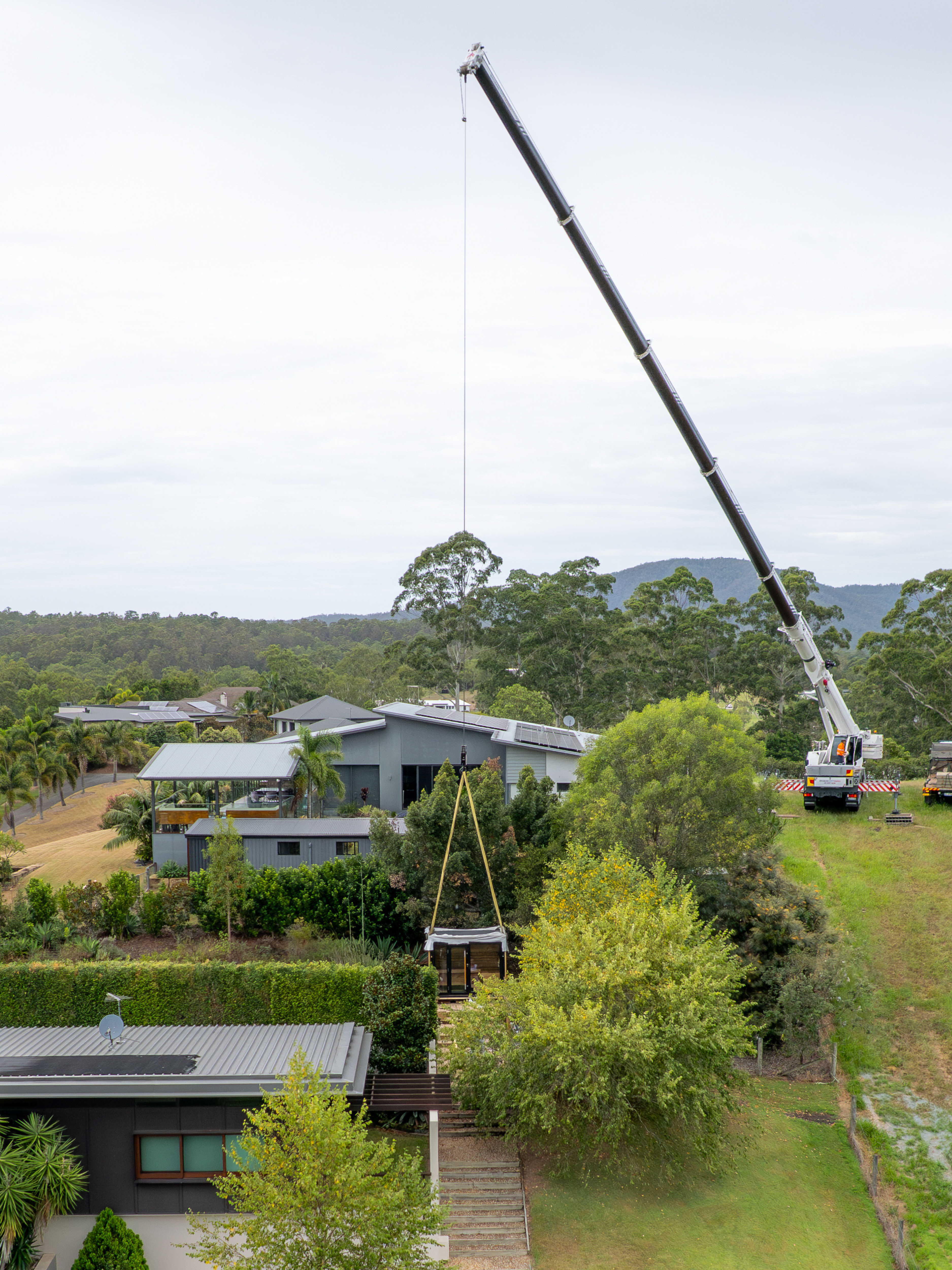  Crane lifting a large object in a rural setting with houses and trees in the background.