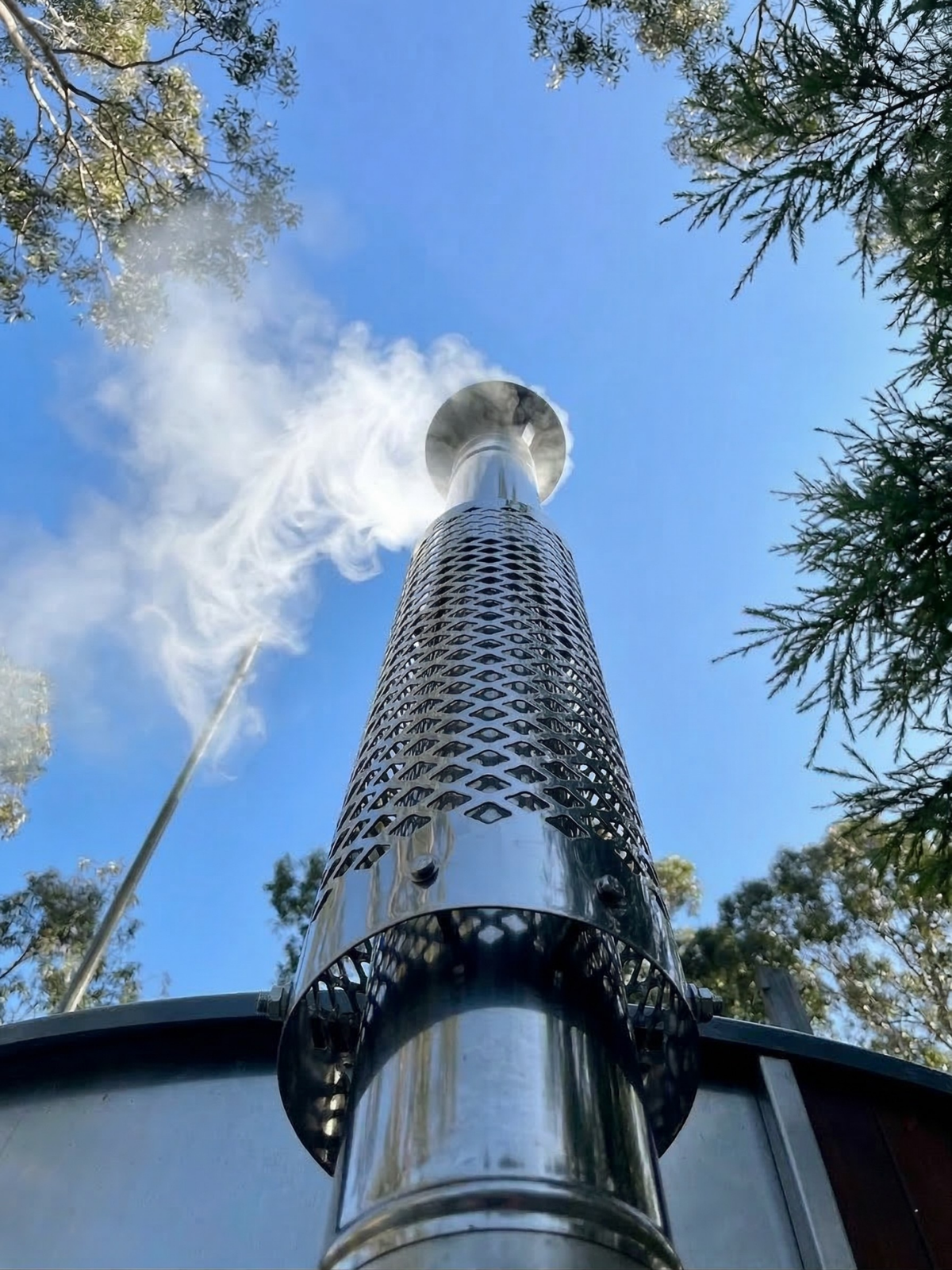 Metal chimney with smoke rising against a blue sky and tree line