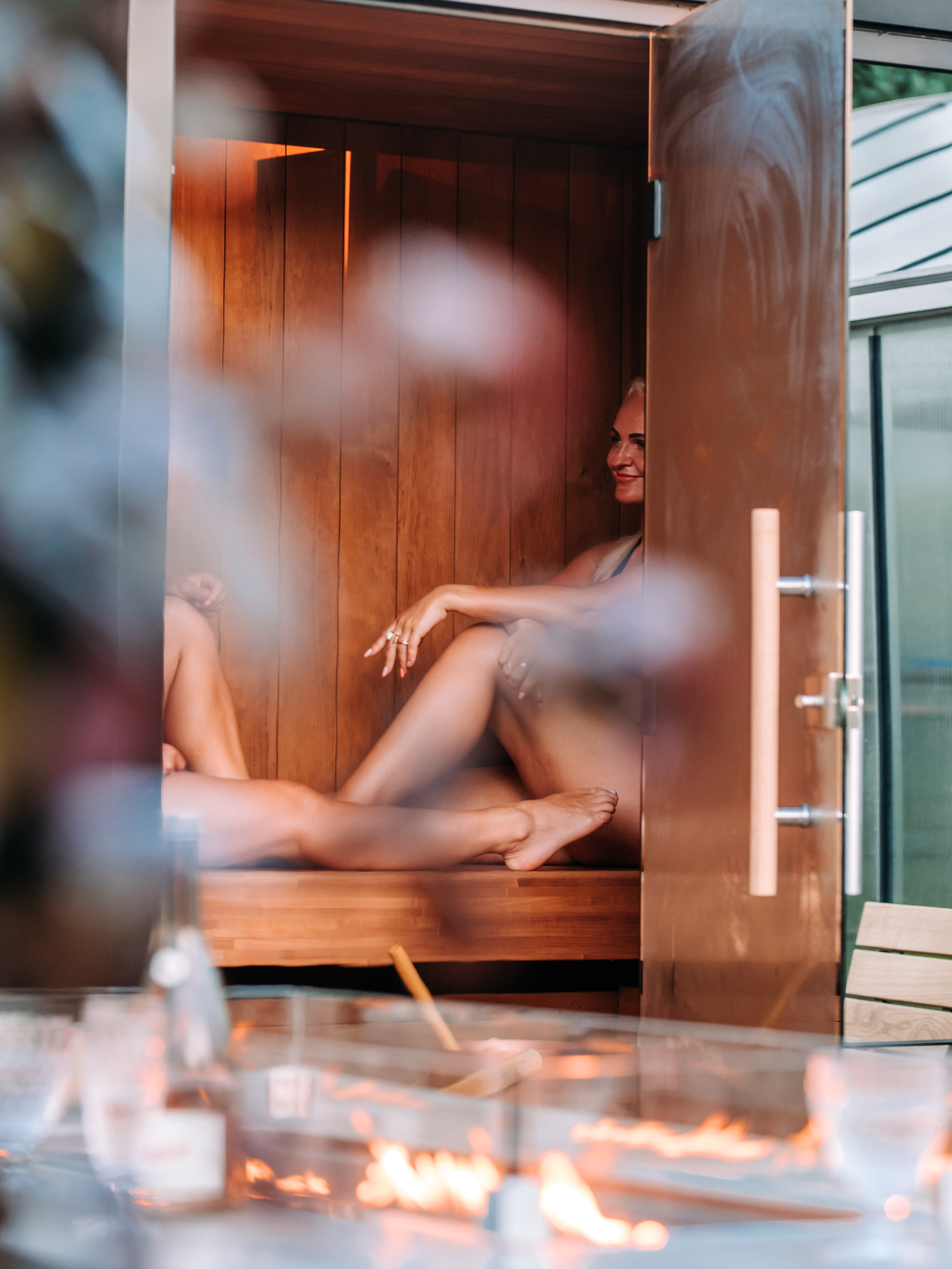 Two women sitting in a wooden sauna with a blurred foreground of flames.