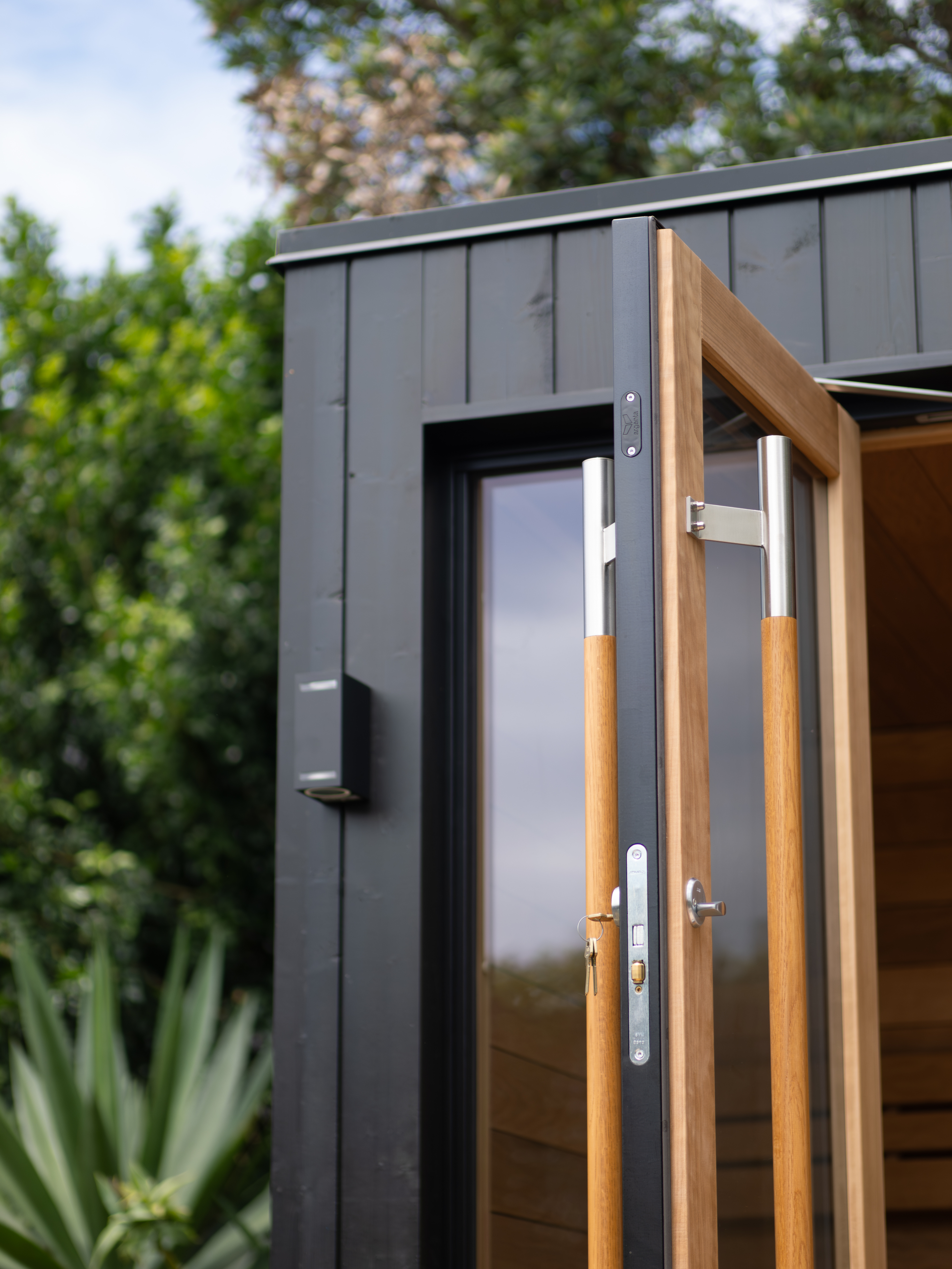 Small wooden cabin with a glass door and black exterior, surrounded by greenery.