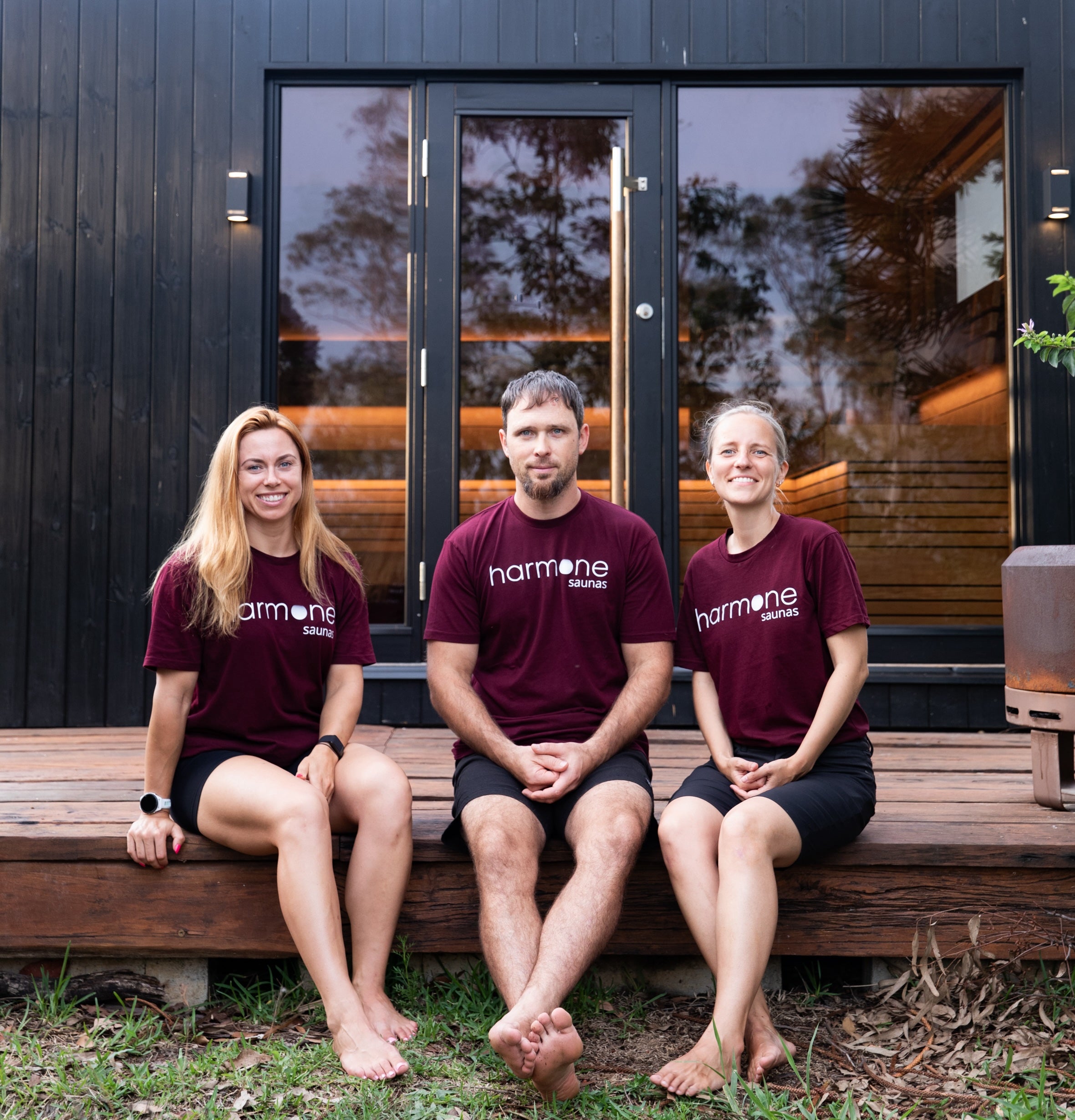 Three people wearing maroon 'harmone' t-shirts sitting on a wooden bench outside a modern building.
