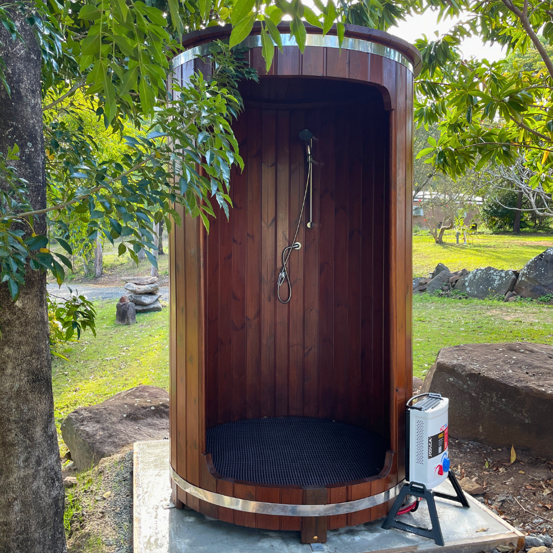 Wooden outdoor sauna with a dark wood finish, set in a serene garden surrounded by lush green trees and grass. A heater is visible outside.
