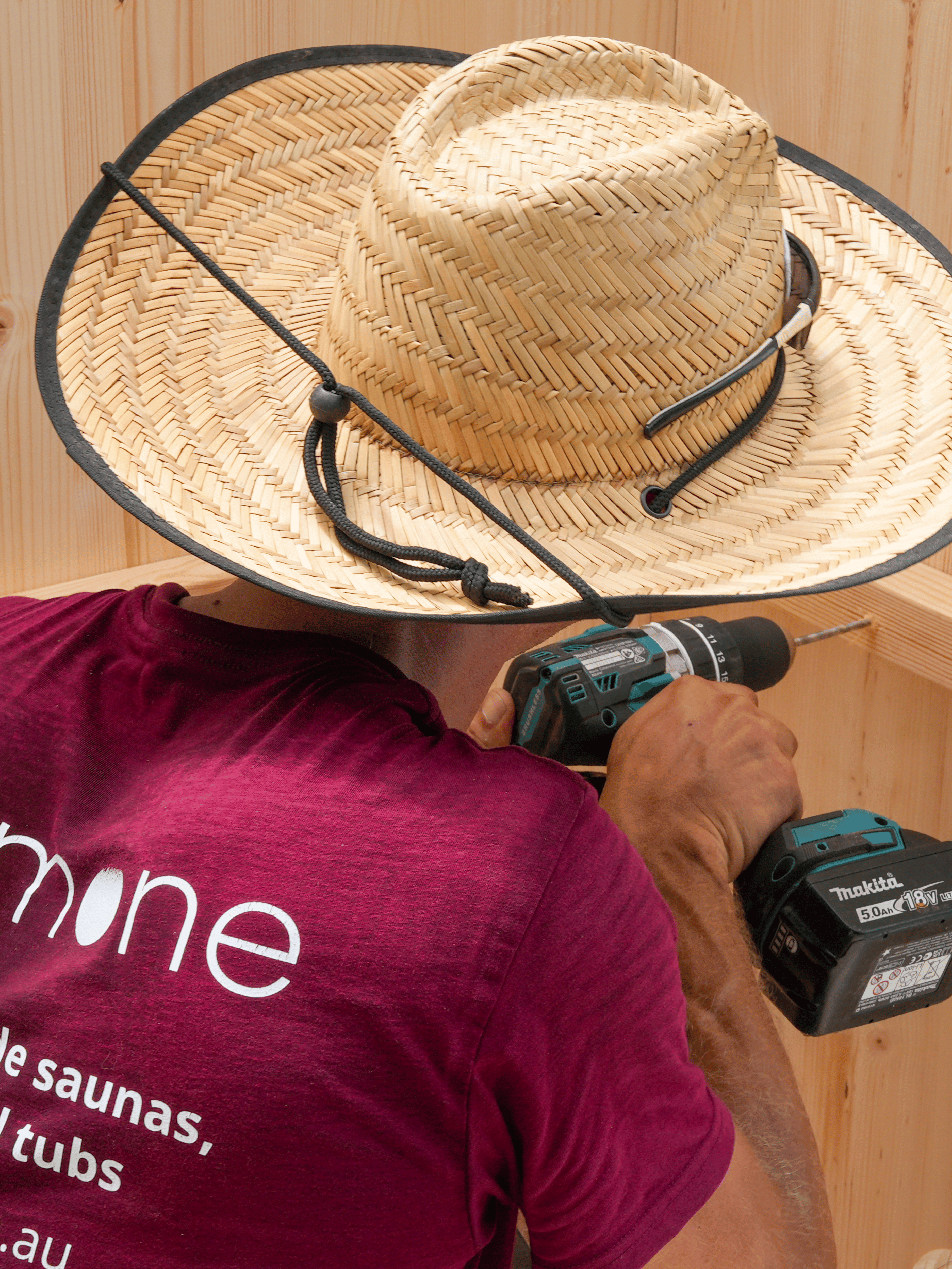Person wearing a straw hat and holding a power drill, with a visible brand logo on a maroon shirt.